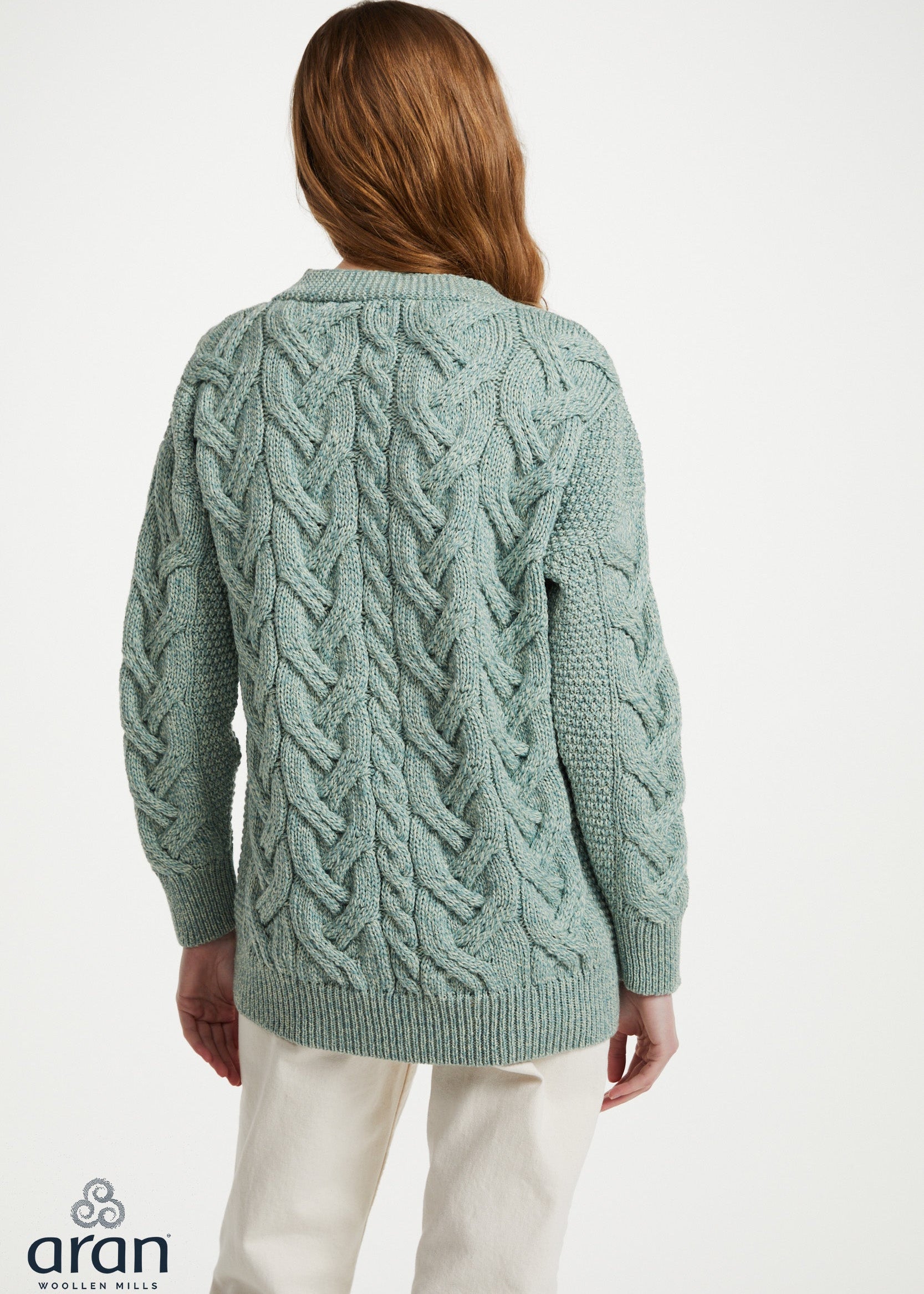 A woman viewed from the back wearing a sage green cable-knit cardigan sweater and cream trousers, against a plain white studio background. The Aran brand logo is visible in the bottom-left corner.