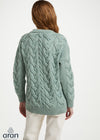 A woman viewed from the back wearing a sage green cable-knit cardigan sweater and cream trousers, against a plain white studio background. The Aran brand logo is visible in the bottom-left corner.