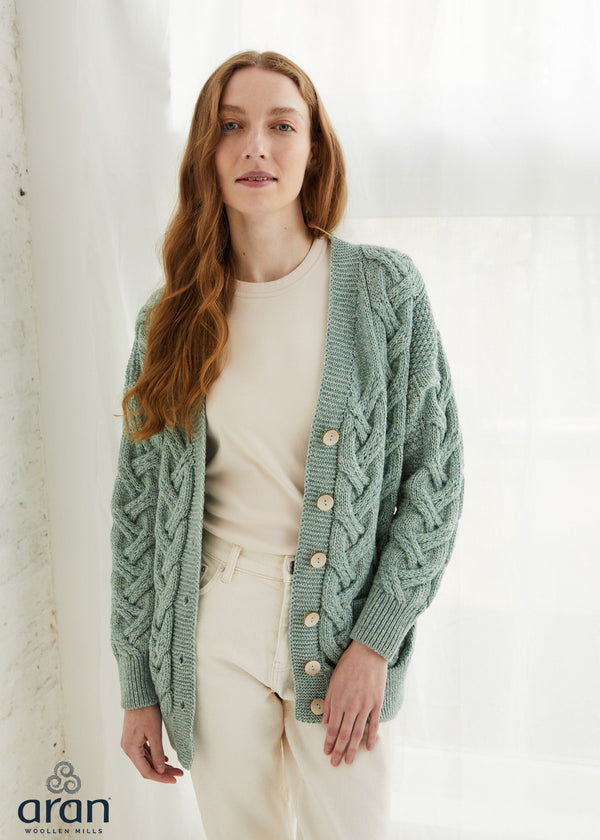 A woman wearing a sage green cable-knit cardigan with wooden buttons over a cream top and cream trousers, standing in front of a white curtain. The Aran Collection logo is visible in the bottom-left corner.