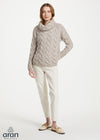 A woman modeling a chunky cable-knit cowl-neck sweater in a light taupe-beige color, paired with cream trousers and beige loafers, against a plain white studio background. The 'aran' brand logo is visible in the bottom-left corner.