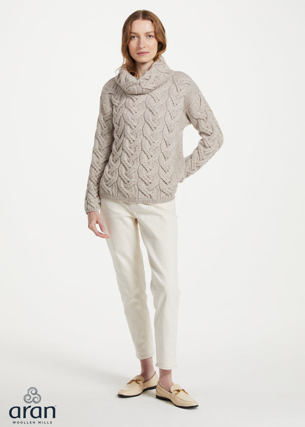 A woman modeling a chunky cable-knit cowl-neck sweater in a light taupe-beige color, paired with cream trousers and beige loafers, against a plain white studio background. The 'aran' brand logo is visible in the bottom-left corner.