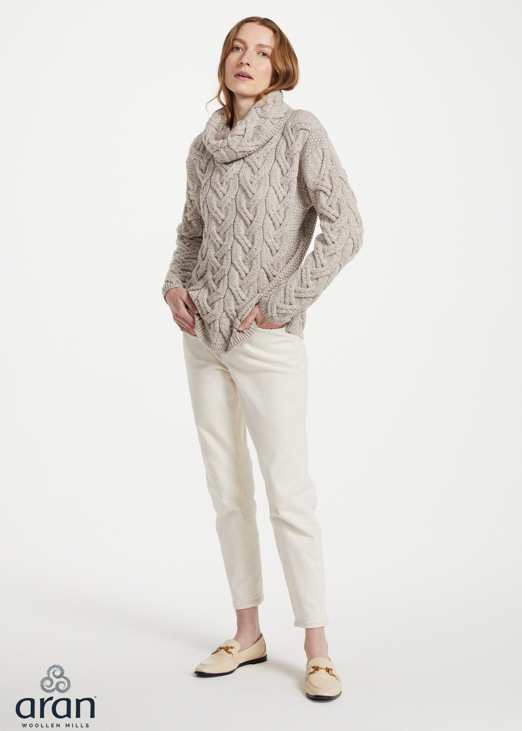 A woman modeling a chunky cable-knit cowl-neck sweater in a light taupe-beige color, paired with off-white trousers and beige loafers, against a plain white studio background. The 'aran' brand logo is visible in the bottom-left corner.