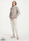 A woman modeling a chunky cable-knit cowl-neck sweater in a light taupe-beige color, paired with off-white trousers and beige loafers, against a plain white studio background. The 'aran' brand logo is visible in the bottom-left corner.
