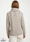 A woman viewed from the back wearing a light oatmeal cable-knit sweater with a high folded collar and white trousers, against a plain light background. The Aran logo is visible in the bottom-left corner.
