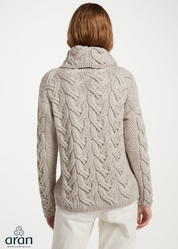A woman viewed from the back wearing a light oatmeal cable-knit sweater with a high folded collar and white trousers, against a plain light background. The Aran logo is visible in the bottom-left corner.