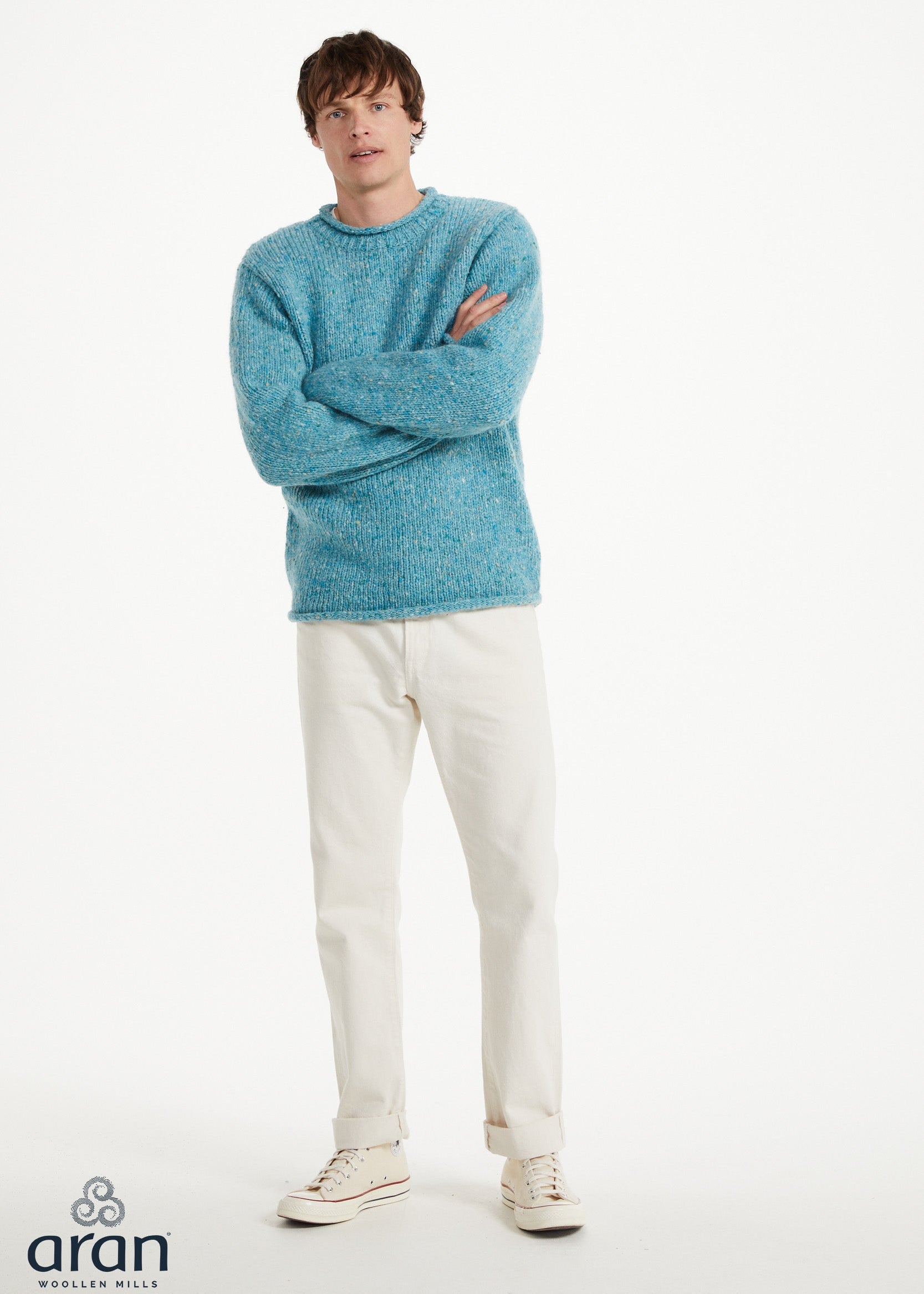 A man modeling a light blue marled knit sweater, paired with off-white trousers and white sneakers, standing against a plain white studio background. The 'aran' brand logo is visible in the bottom-left corner.