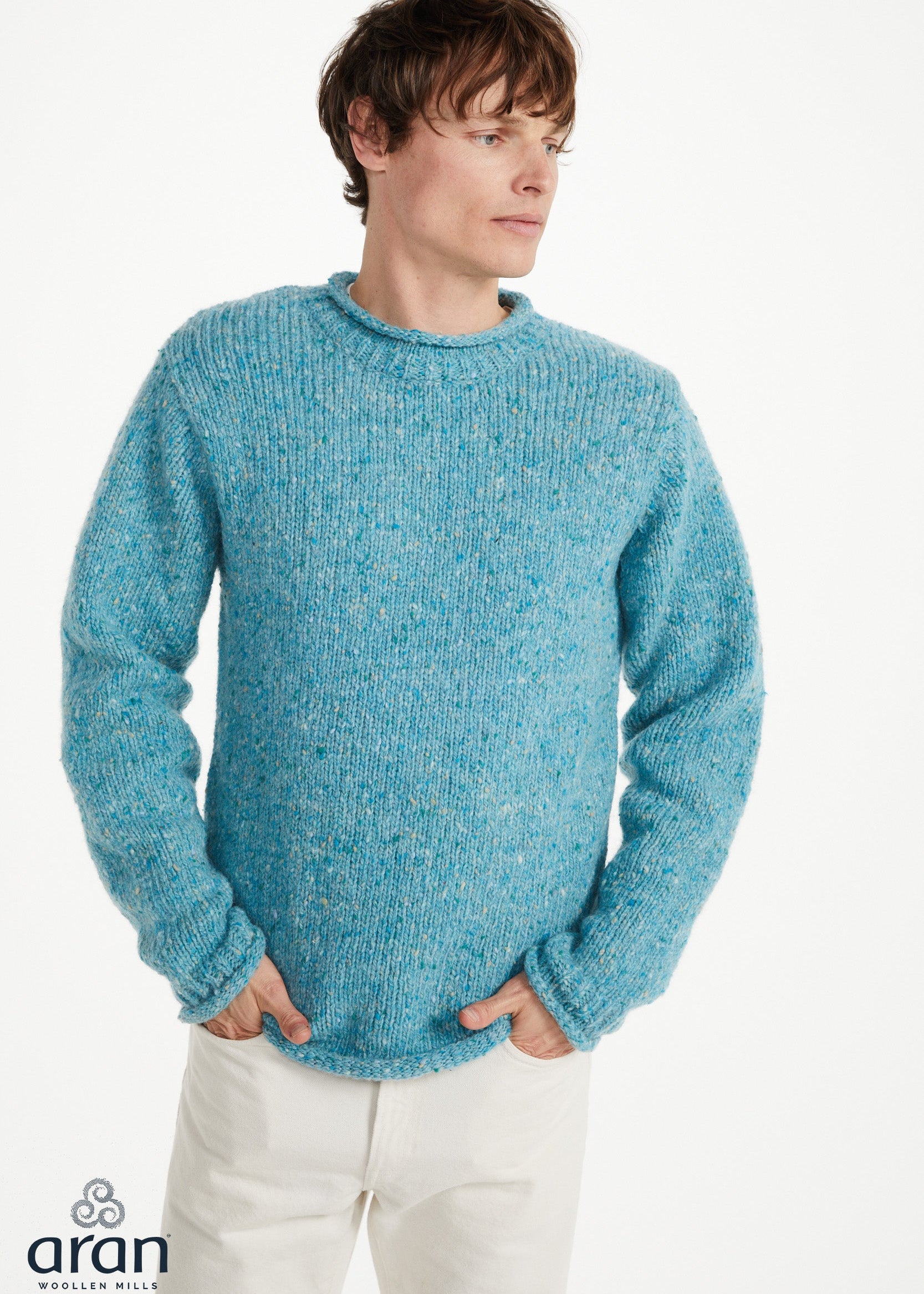 A man wearing a textured, chunky knit crewneck sweater in a heathered light blue color, paired with off-white trousers, against a plain white studio background. The Aran logo is visible in the bottom-left corner.
