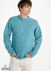 A man wearing a textured, chunky knit crewneck sweater in a heathered light blue color, paired with off-white trousers, against a plain white studio background. The Aran logo is visible in the bottom-left corner.