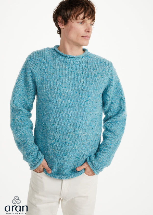 A man wearing a textured, chunky knit crewneck sweater in a heathered light blue color, paired with off-white trousers, against a plain white studio background. The Aran logo is visible in the bottom-left corner.