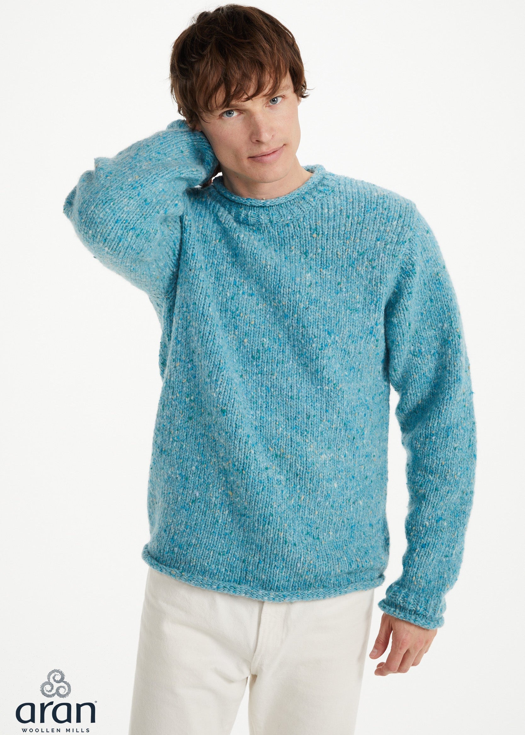 A man wearing a textured, heathered light blue crewneck sweater and cream trousers, posing against a plain white studio background. The 'aran' brand logo is visible in the bottom-left corner.