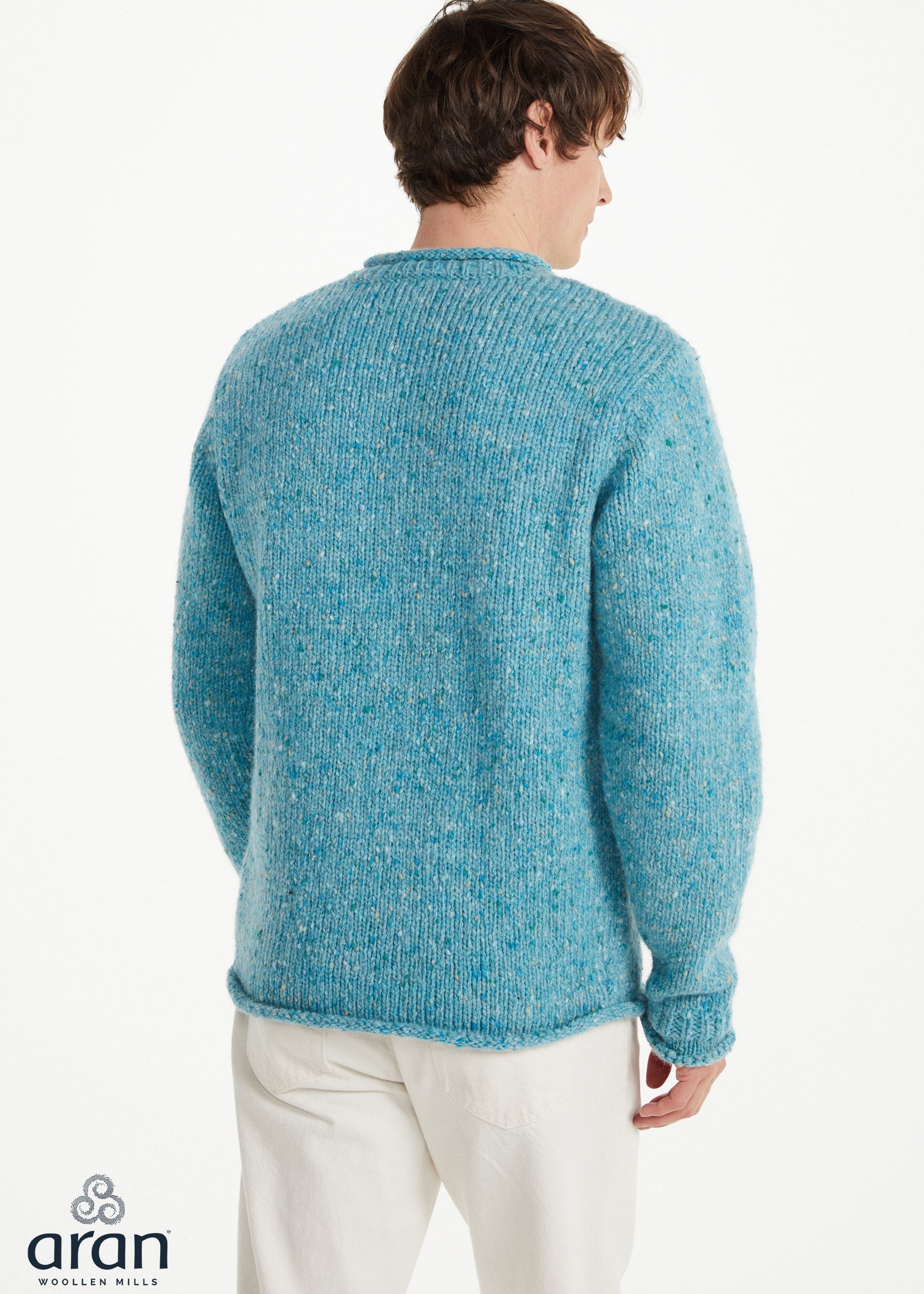 A man viewed from behind wearing a textured, heathered blue knit sweater and cream trousers, against a plain white studio background. The 'aran woolen mills' logo is visible in the bottom-left corner.