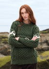 A woman with red hair wearing a dark green Aran cable-knit sweater with white geometric patterns on the chest and sleeves, standing outdoors with her arms crossed. The Aran Crafts logo is visible in the bottom right corner.