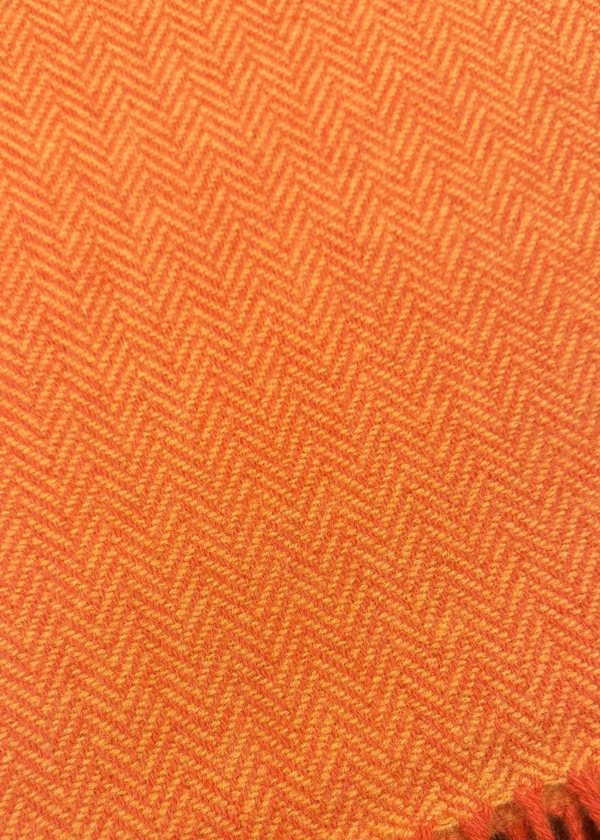 A close-up of an orange woven textile with a herringbone pattern and fringed edge, laid on a dark surface.