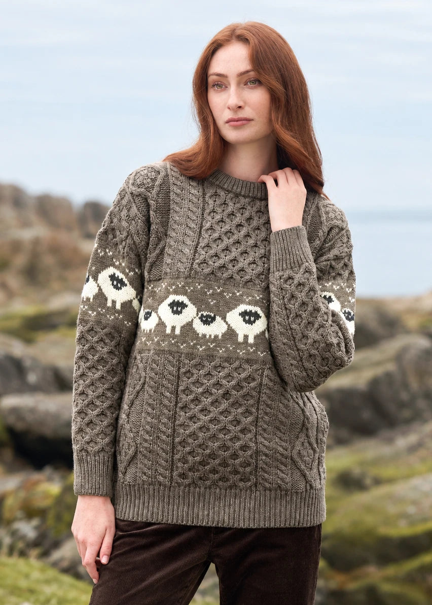 A woman wearing a chunky Aran-style cable-knit sweater with a horizontal band of white sheep motifs in the center, photographed outdoors against a blurred rocky coastal background. The sweater is a warm taupe/gray-brown color, and the sheep pattern is white with black faces.