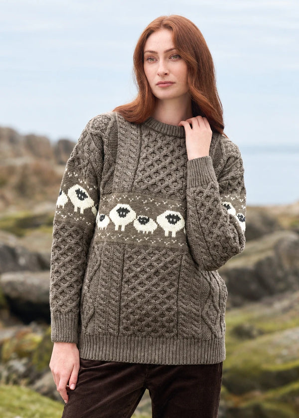 A woman wearing a chunky Aran-style cable-knit sweater with a horizontal band of white sheep motifs in the center, photographed outdoors against a blurred rocky coastal background. The sweater is a warm taupe/gray-brown color, and the sheep pattern is white with black faces.