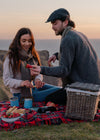 A couple enjoys a picnic on a grassy cliffside overlooking the ocean at sunset. They sit on a red and black plaid blanket with a wicker basket, blue mugs, and food, while the man offers a strawberry to the woman.
