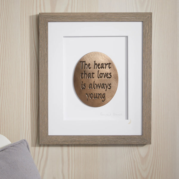Framed wooden plaque with 'The heart that loves is always young' quote on a wooden wall.