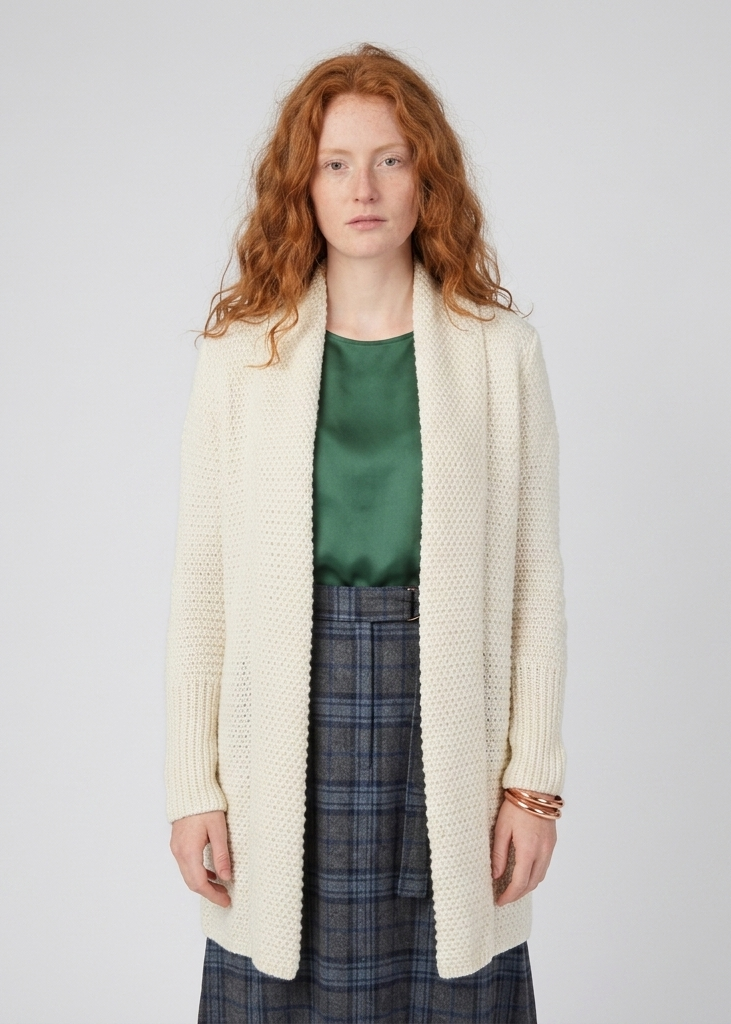 A model with red hair wearing a cream-colored, open-front, long-sleeve knit cardigan over a green satin top and a blue plaid skirt, standing against a light gray studio background.