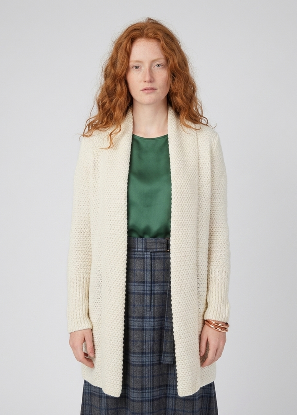 A model with red hair wearing a cream-colored, open-front, long-sleeve knit cardigan over a green satin top and a blue plaid skirt, standing against a light gray studio background.