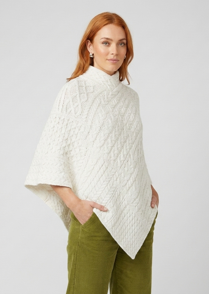A woman modeling a white cable-knit mock-neck poncho sweater, paired with olive green pants, against a light gray studio background.