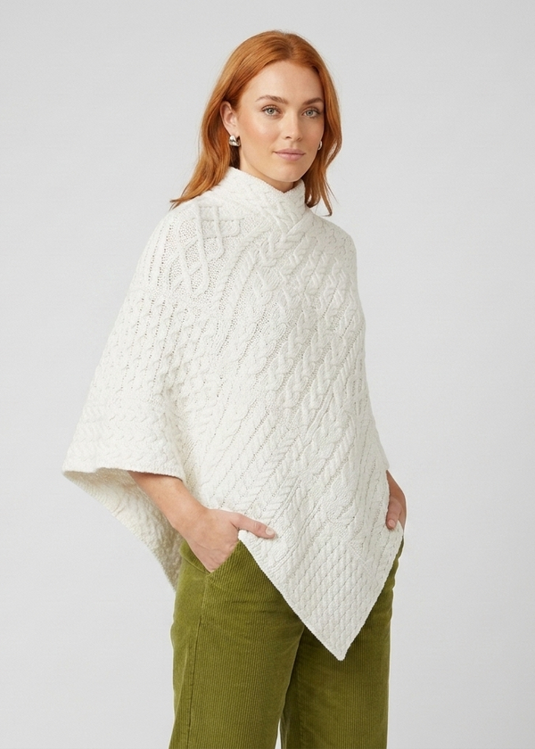 A woman modeling a white cable-knit mock-neck poncho sweater, paired with olive green pants, against a light gray studio background.