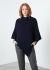 A woman with red hair wearing a dark navy blue cable-knit poncho sweater over a gray long-sleeve shirt and gray trousers, standing in a minimalist white studio setting.