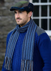 Man wearing a blue knit sweater, scarf, and cap against a stone wall.