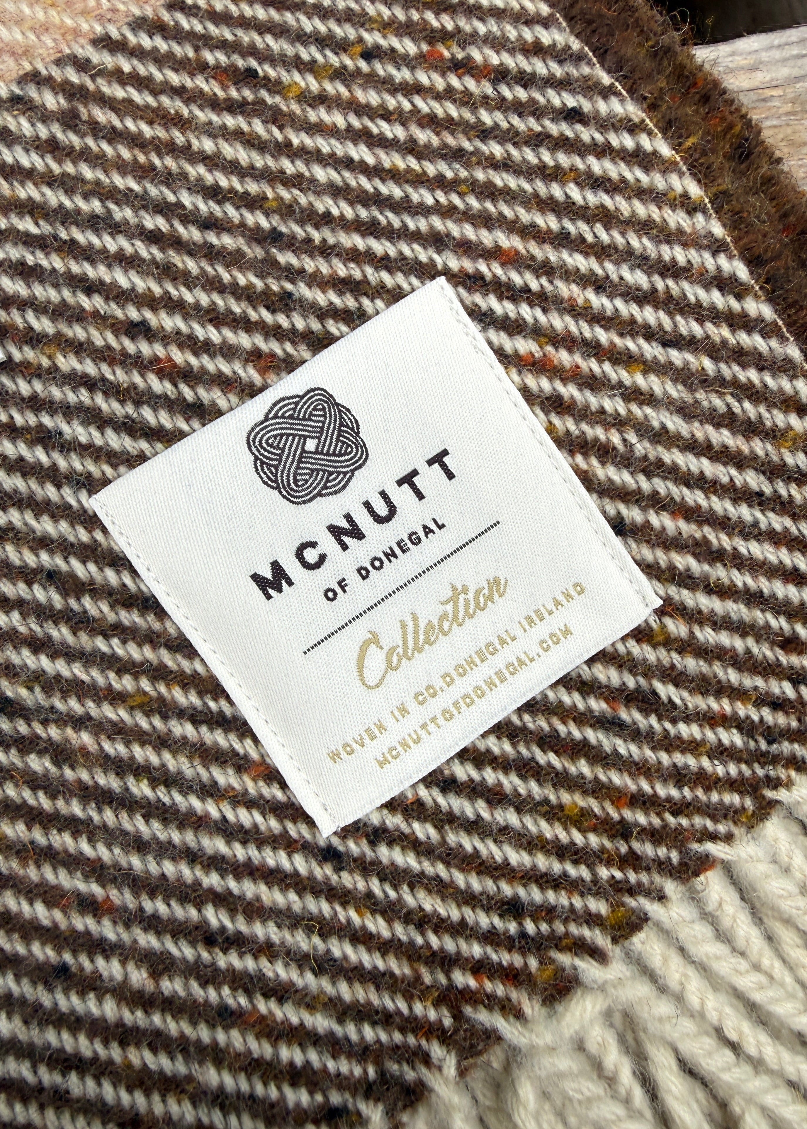 Close-up of a woven wool label reading 'MCNUTT OF DOMEVAL Collection' placed on a brown and cream herringbone textile, with part of a cream knit edge visible at the bottom right.