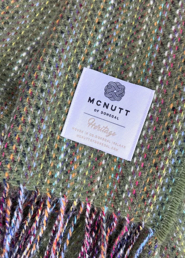 A close-up of a woven blanket or throw in olive green with multicolored yarn flecks and fringed edges, featuring a white McNutt of Donegal Heritage label.