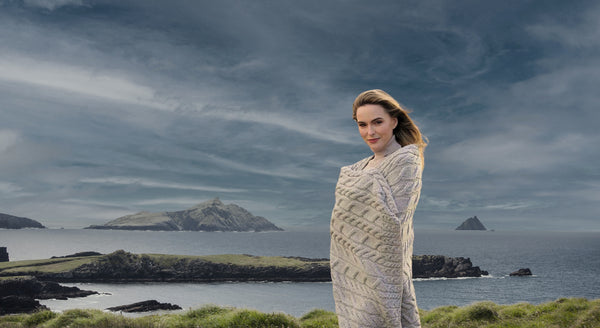 Woman wrapped in a patterned shawl standing on a coastal landscape with islands and a dark sky.