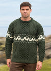 A man wearing a dark green Aran-style cable-knit sweater with a cream sheep pattern across the chest and sleeves, standing outdoors with a blurred coastal background. The Aran Crafts Ireland logo is visible in the bottom right corner.