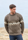 A man wearing a brown and beige Aran-style cable-knit sweater with a sheep pattern across the chest, standing outdoors against a soft-focus background.