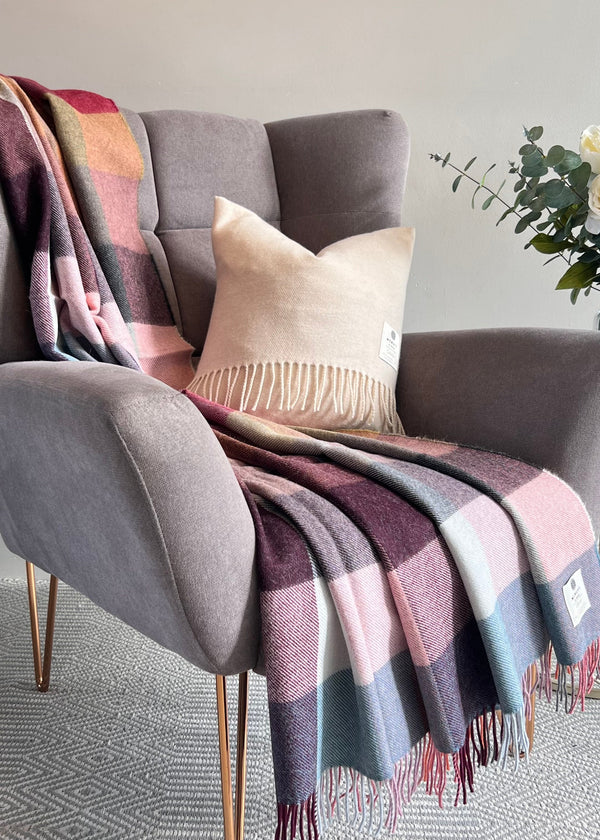 A gray upholstered armchair in a styled room setting, draped with a multicolor plaid throw blanket and a cream fringed pillow, with a side table and flowers in the background.