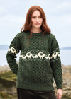A woman wearing a dark green Aran-style knit sweater with a cream sheep pattern across the chest and sleeves, photographed outdoors against a soft-focus coastal background. The Aran Crafts logo is visible in the bottom right corner.