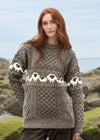 A woman with long red hair wearing a gray cable-knit sweater with a cream and black sheep pattern across the chest and sleeves, standing outdoors against a blurred coastal background. The Aran Crafts logo is visible in the bottom right corner.