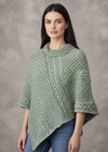 A woman with wavy red hair models a sage green cable-knit poncho sweater against a textured gray studio background.