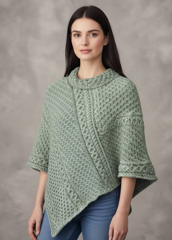 A woman with wavy red hair models a sage green cable-knit poncho sweater against a textured gray studio background.