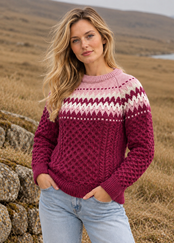 A woman wearing a burgundy and pink Fair Isle cable-knit sweater with light blue jeans, standing outdoors in a grassy, hilly landscape.