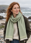 A woman with red hair stands on a rocky beach wearing a light sage green knit foxford scarf with cream fringe, a cream knit sweater, and a dark brown jacket, with the ocean and hills in the background.