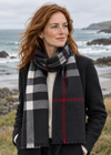 A woman with red hair stands on a beach wearing a dark charcoal coat and a large black, gray, and white plaid foxford scarf with a thin red stripe. The background shows the ocean, rocks, and grassy shoreline under an overcast sky.