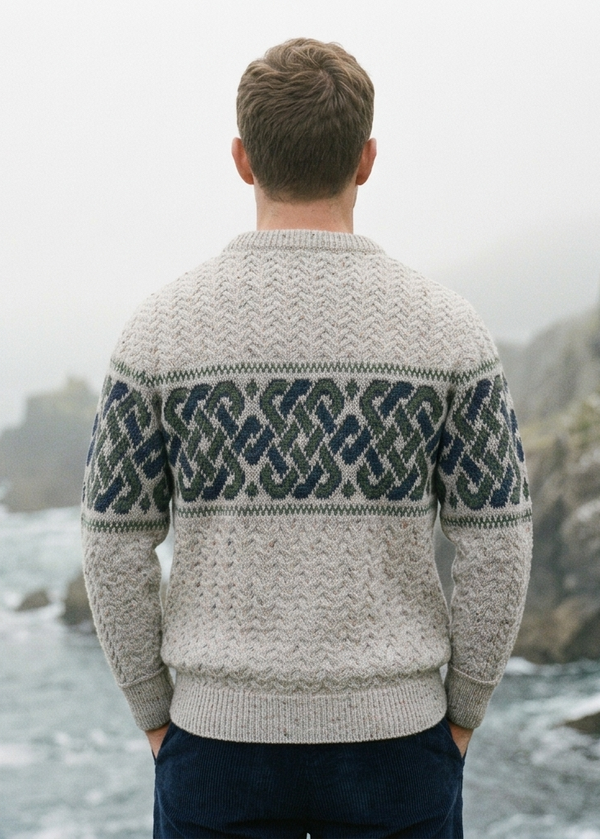 A man viewed from behind wearing a cream-colored cable-knit sweater with a dark navy blue geometric pattern across the shoulders and upper back, standing on a rocky coast with the sea in the background.