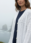 Woman wearing a white textured cardigan over a navy dress with a blurred coastal background