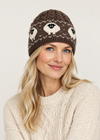 A woman wearing a brown cable-knit beanie with a cream and black Nordic eye pattern, paired with a cream cable-knit sweater, against a light beige background.