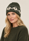 A woman wearing a dark olive green cable-knit beanie with a cream and black Nordic-style pattern, paired with a matching dark green knit sweater, against a solid light beige background.