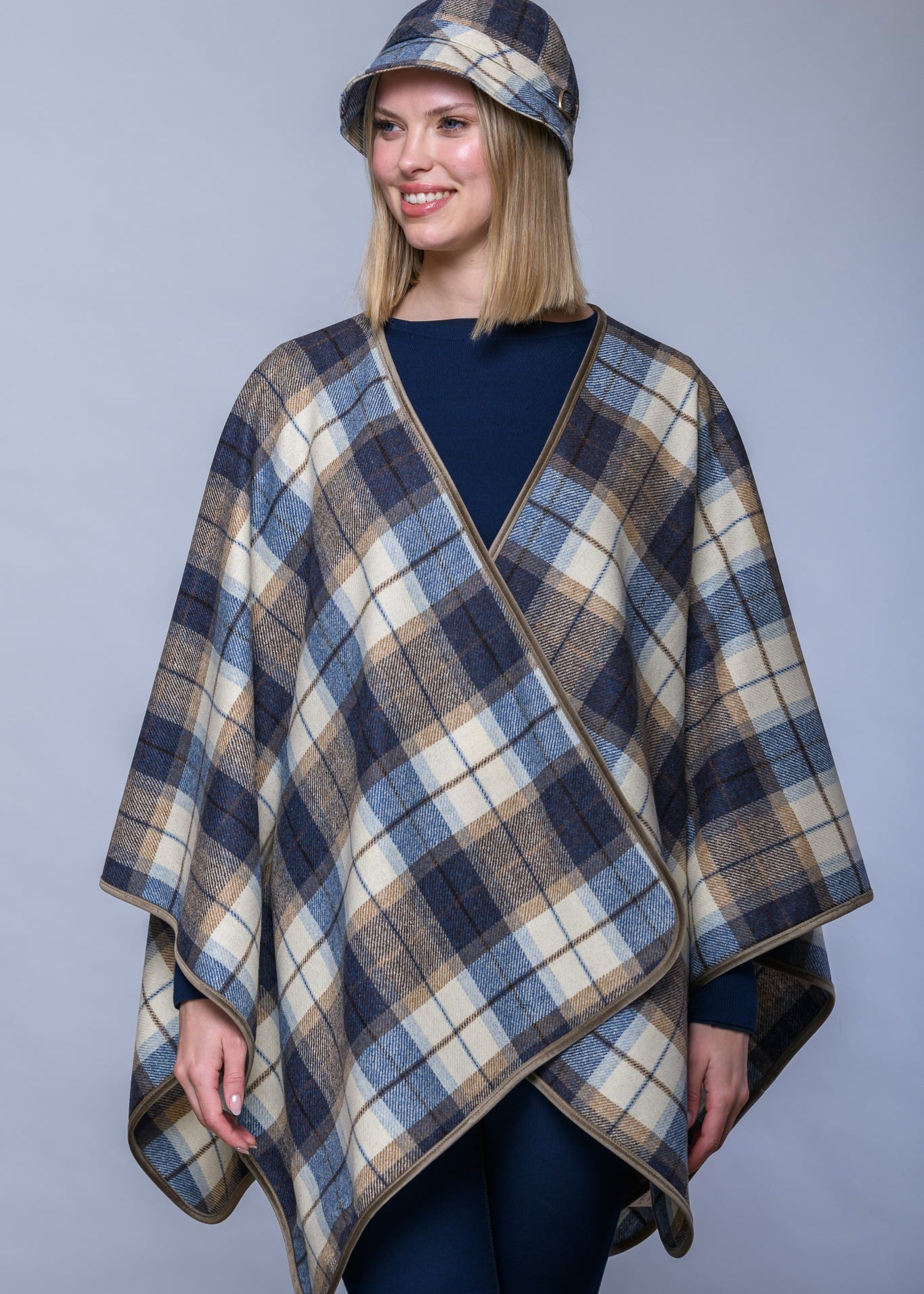 A woman modeling a plaid wrap poncho and matching bucket hat in navy, beige, and brown tones against a solid gray studio background.