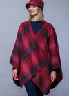 A woman wearing a red and dark green plaid wrap poncho with a matching red plaid hat, photographed against a solid gray studio background.