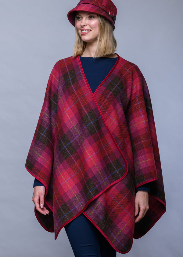 A woman wearing a red and dark green plaid wrap poncho with a matching red plaid hat, photographed against a solid gray studio background.