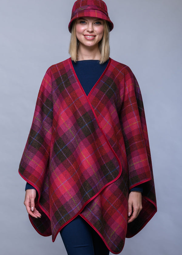 A woman modeling a red and dark green plaid wrap poncho with a matching plaid bucket hat, photographed against a solid gray studio background.