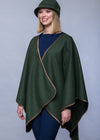 A woman modeling a dark green wrap poncho with tan trim, paired with a matching green hat, navy top, and navy pants, against a solid gray studio background.
