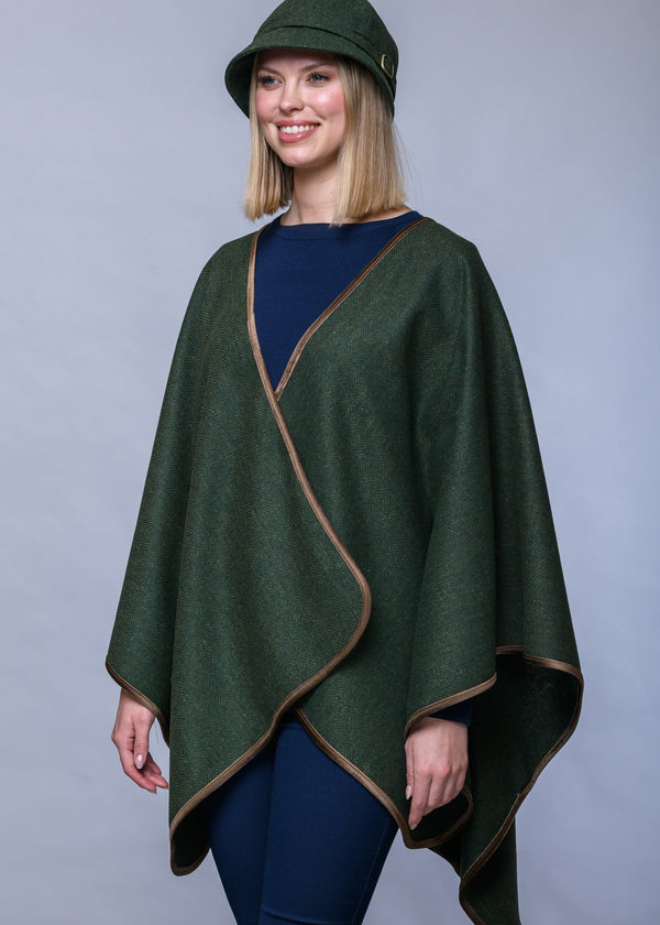 A woman modeling a dark green wrap poncho with tan trim, paired with a matching green hat, navy top, and navy pants, against a solid gray studio background.